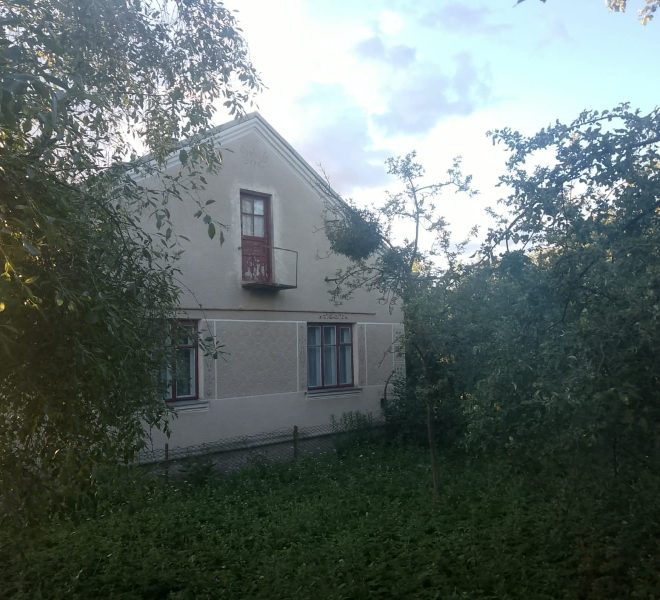 Two-story beige house partially hidden by dense trees, with a small balcony and red door on the upper level.