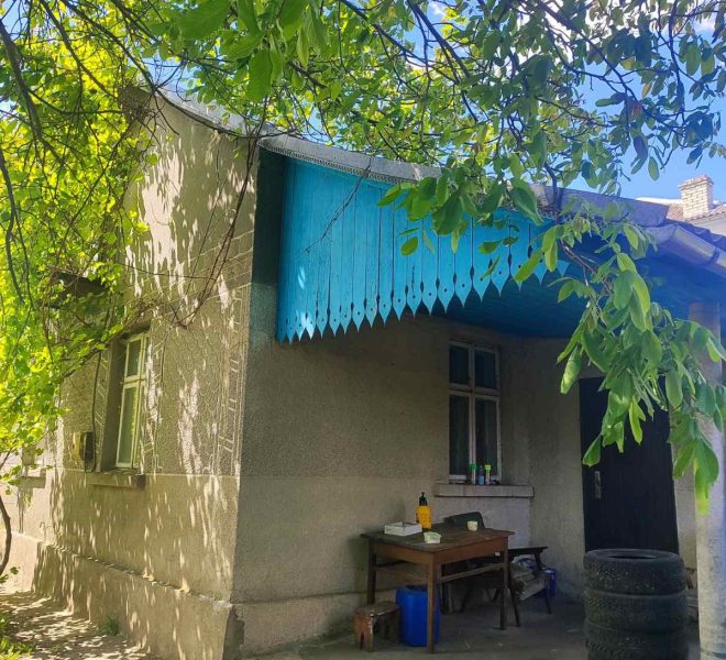 Side of a beige house with a bright blue scalloped awning, a small outdoor work table, and leafy shade from trees above.