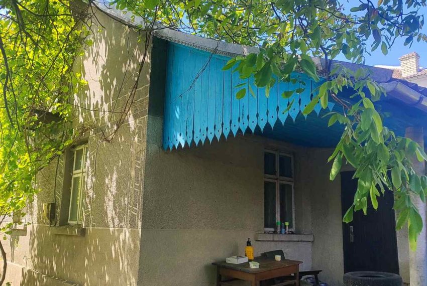 Side of a beige house with a bright blue scalloped awning, a small outdoor work table, and leafy shade from trees above.