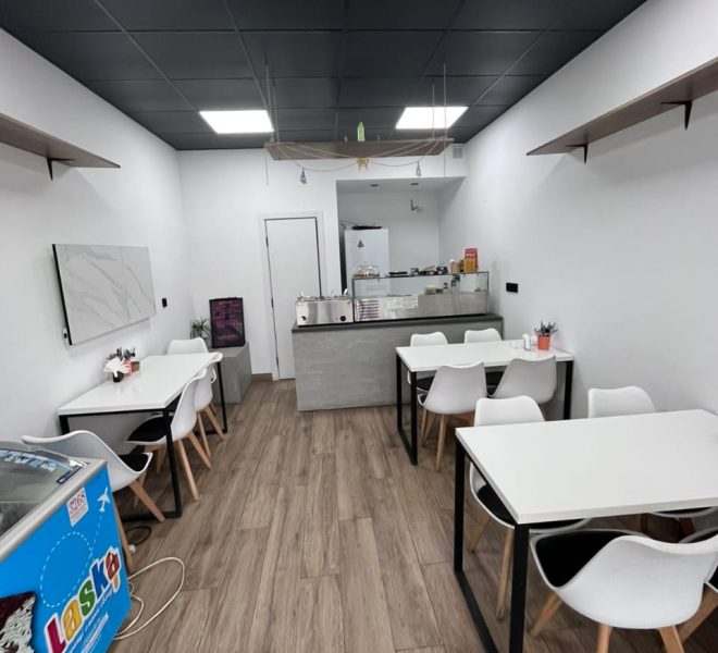 Modern cafe interior with white tables and chairs, wooden floor, and a service counter at the back.