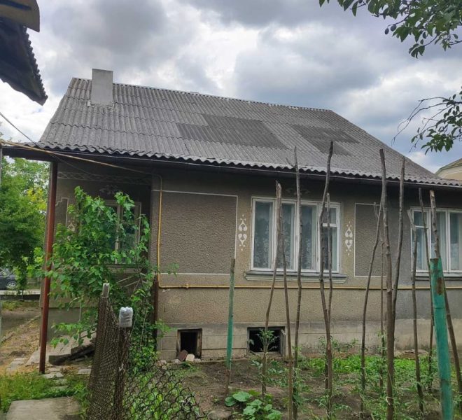 Single-story house with a gray tiled roof, a small garden with vines and wooden stakes, under a cloudy sky.